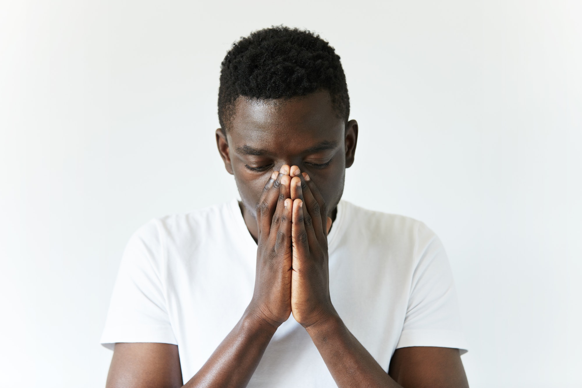 Close up shot of African man wearing white T-shirt standing with depressed and sad look, covering hi